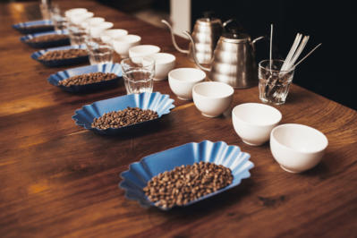 Wooden table top with neat rows of cups, water glasses and blue containers with roated coffee beans laid out in preparation for a professional coffee tasting